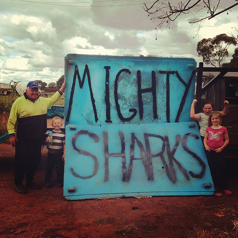 Group of children and adult holding a "Mighty Sharks" sign outdoors during a rugby event.